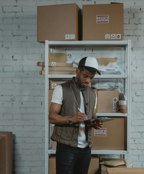 Courier in a warehouse checking inventory on a tablet amidst stacked cardboard boxes.